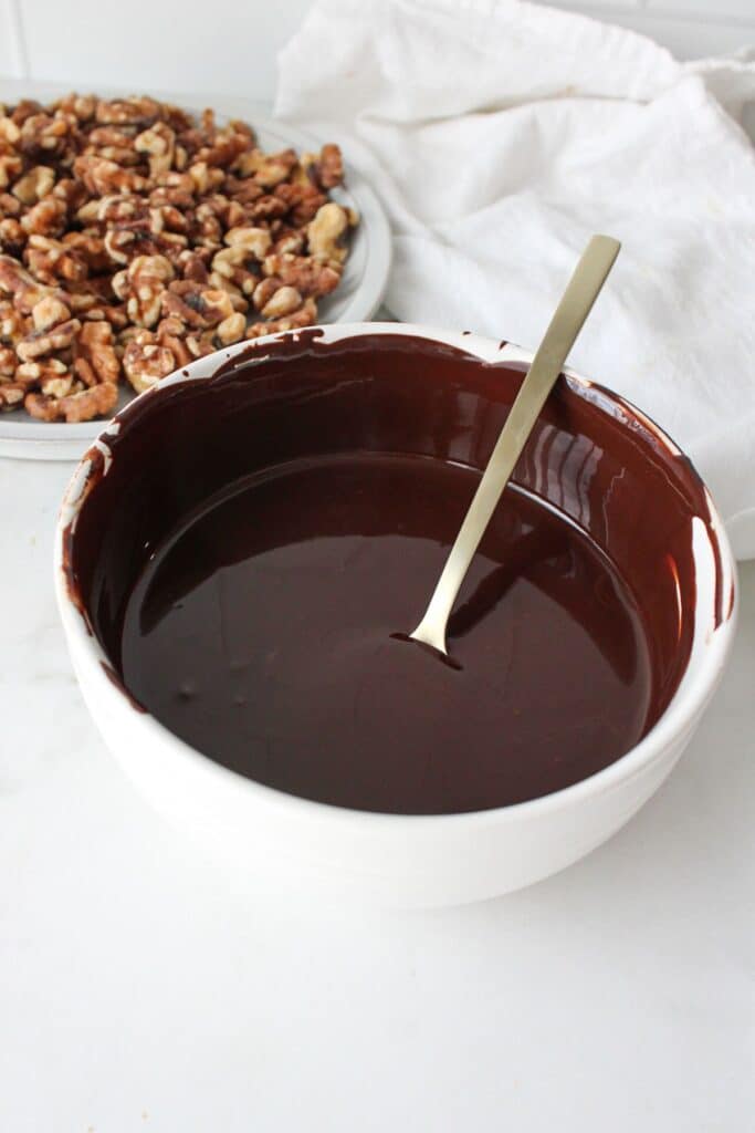 A white bowl of melted dark chocolate with walnuts in the background.
