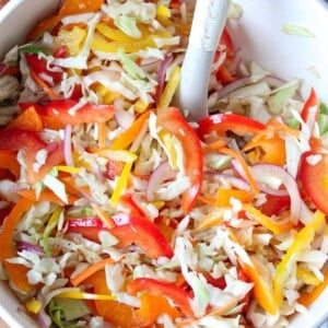 A close up shot of bell pepper slaw in a white bowl with a serving spoon.