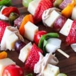 A close up photo of charcuterie skewers on a wooden board with various cheese, fruit and meat.