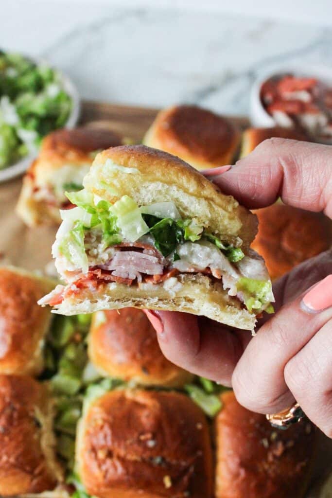 A hand holding up a chopped italian sliders with many sliders in the background.