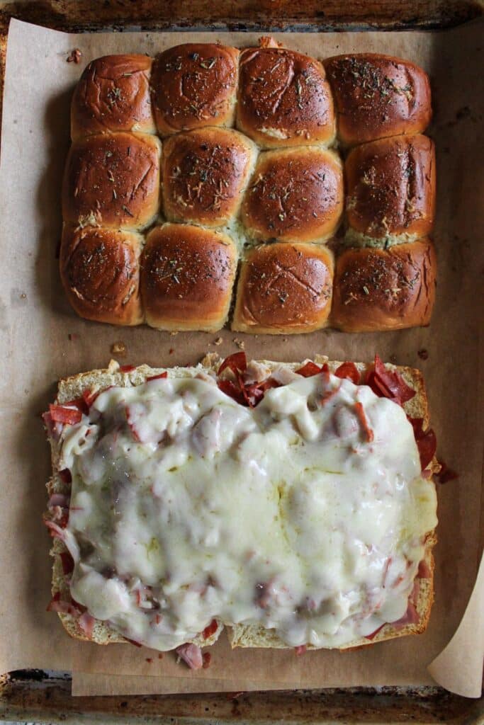 Toasted slider buns on a sheet pan with meats and melted cheese on a sheet pan.