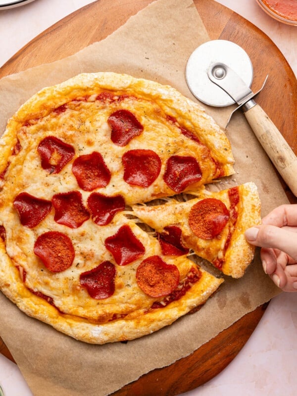 A hand removes a slice of pizza from the cottage cheese pizza sitting on a piece of parchment paper on top of a wooden cutting board.