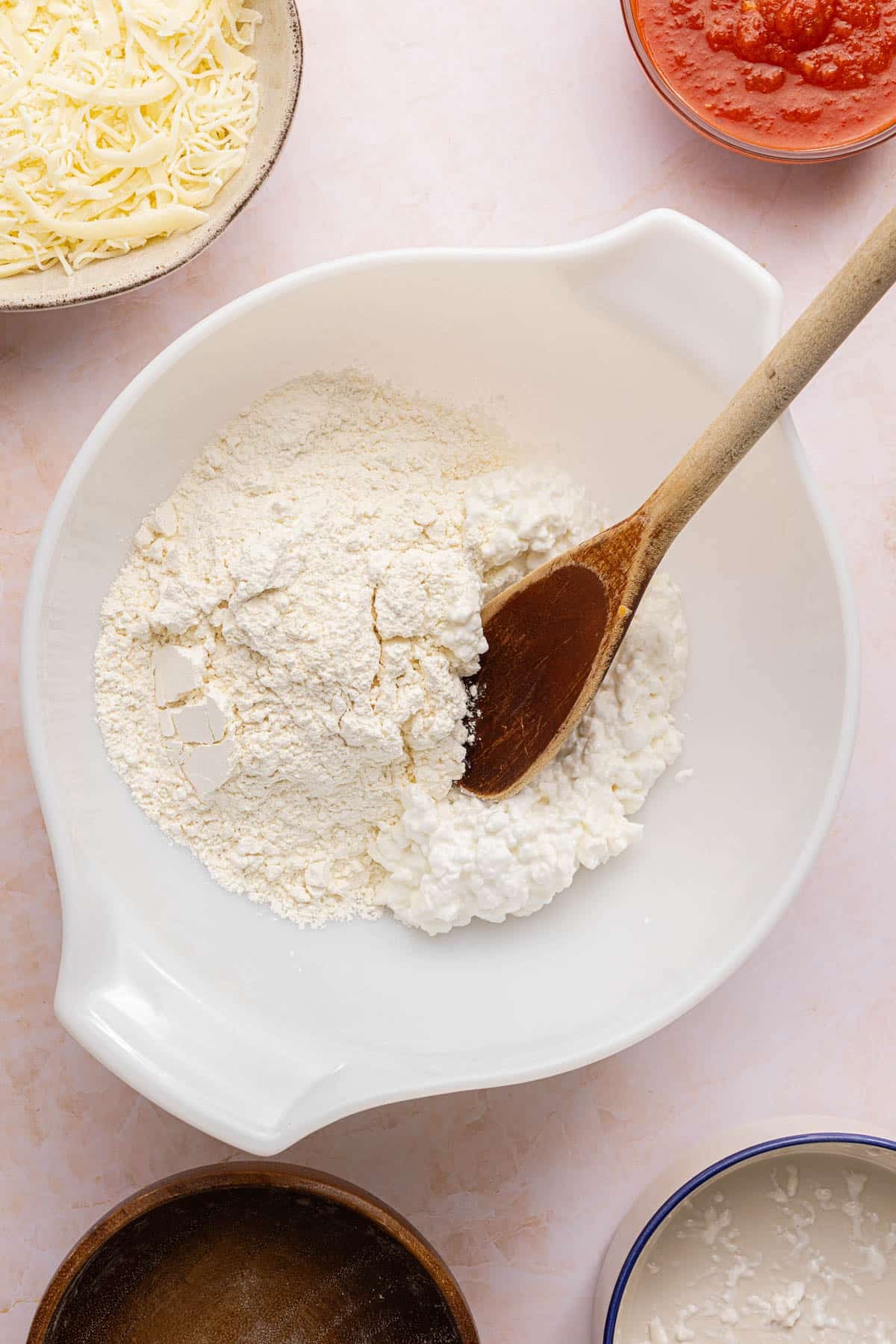 The self-rising flour and cottage cheese in a white bowl on a countertop with a wooden spoon in it.