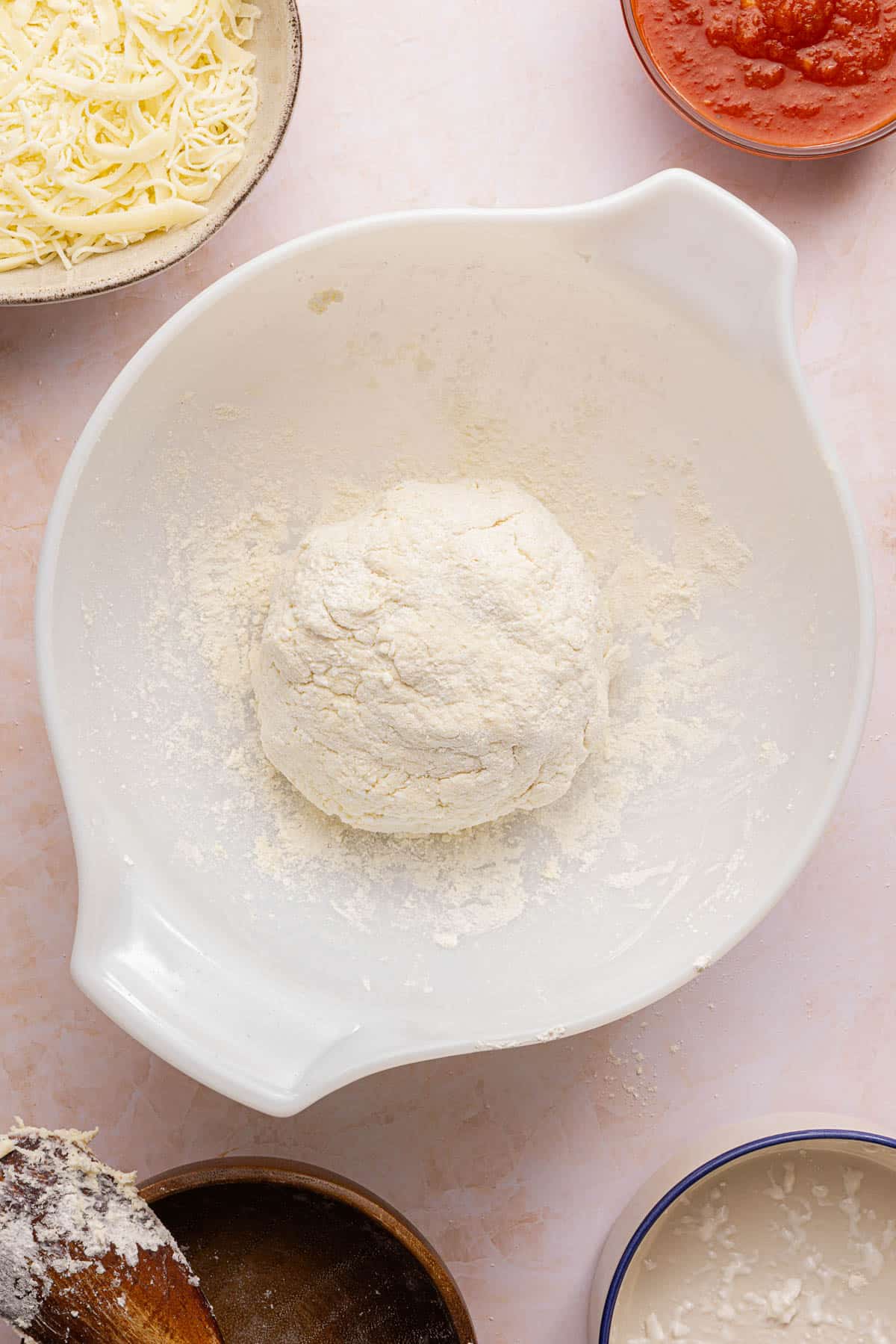 The pizza dough formed into a ball in a white bowl on a countertop.