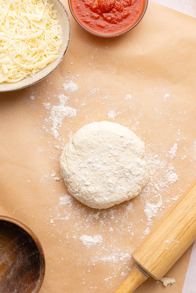 The pizza dough ball on a piece of brown parchment paper sprinkled with flour. A rolling pin sits off to the right side.