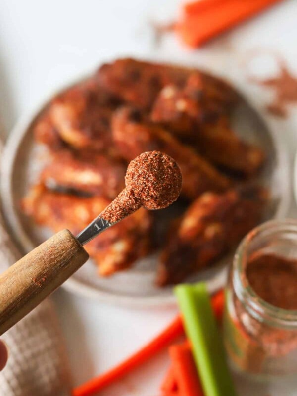 A close up shot of buffalo chicken dry rub in a measuring spoon above a plate of buffalo chicken wings.