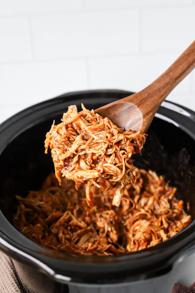 Brown sugar BBQ chicken being scooped out of a crock pot with a wooden spoon.