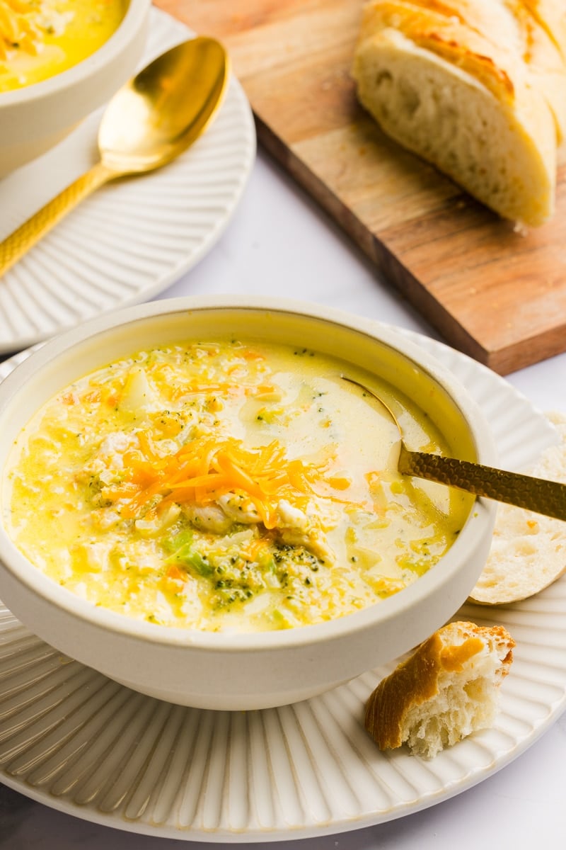 A bowl of chicken broccoli cheddar soup in a white bowl with some bread.
