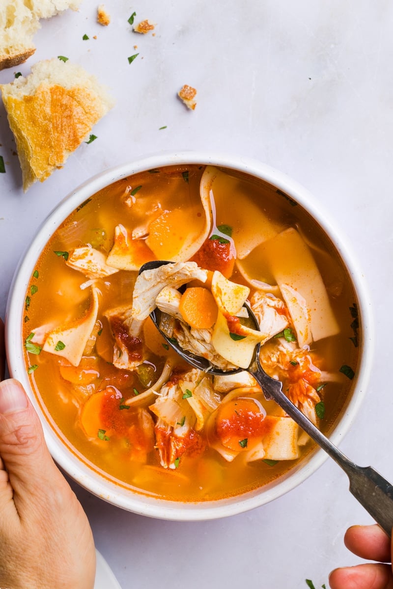 Spicy chicken noodle soup with sriracha in a white bowl with a hand taking a spoonful.