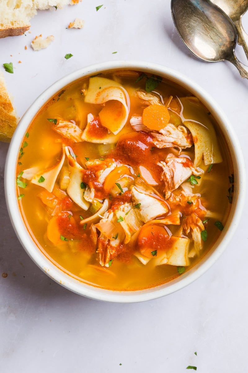 Spicy chicken noodle soup topped with sriracha in a white bowl.