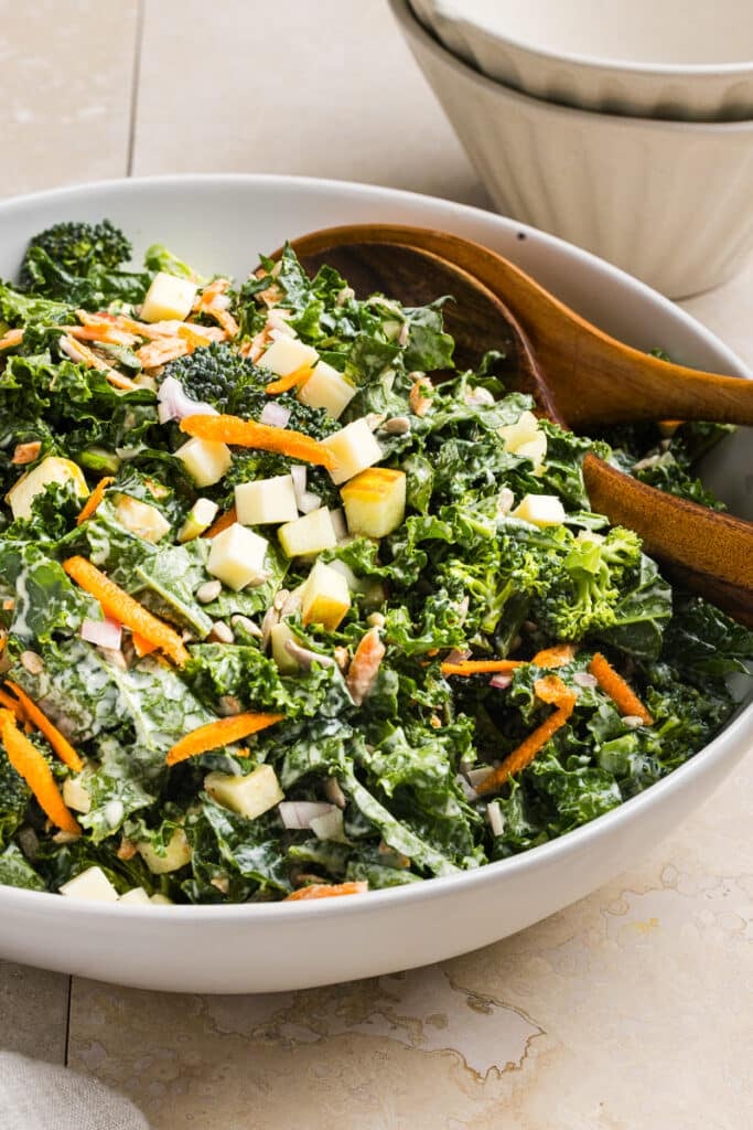 A close up shot of broccoli and kale salad in a bowl tossed with dressing and two wooden spoons.