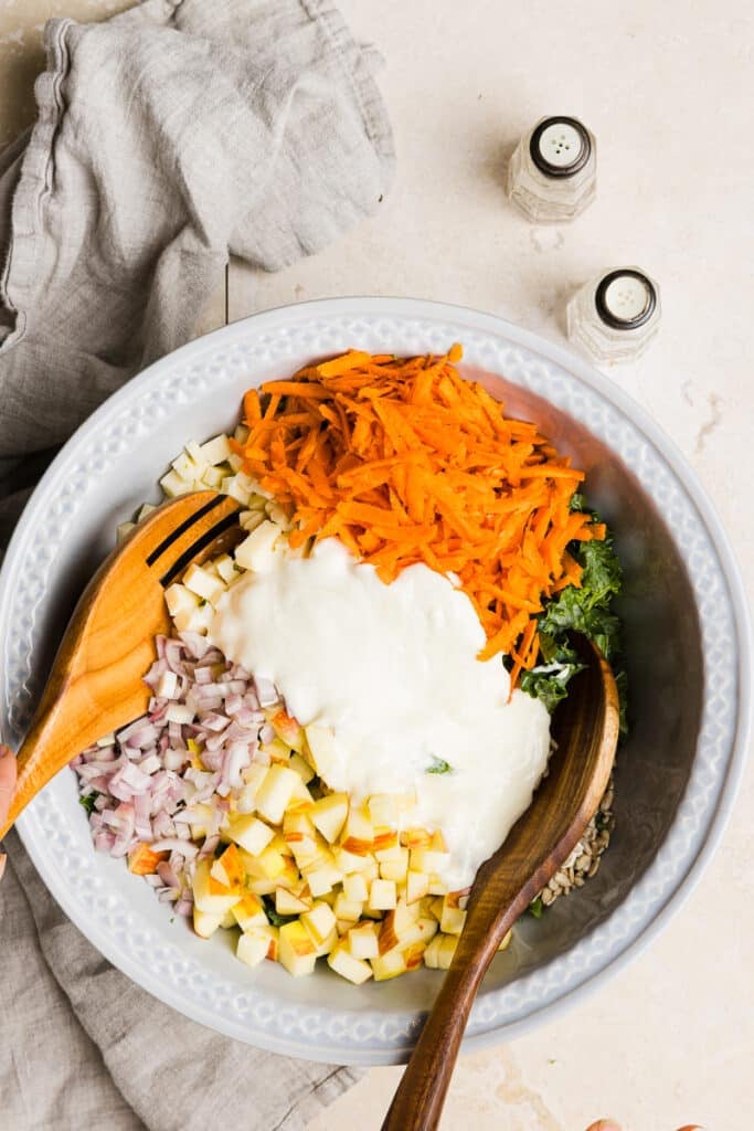 Greek yogurt dressing being added to kale and broccoli salad in a large bowl with two wooden spoons.
