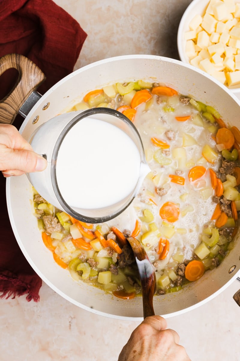 Milk being poured into a large pot with veggies, potatoes and broth.