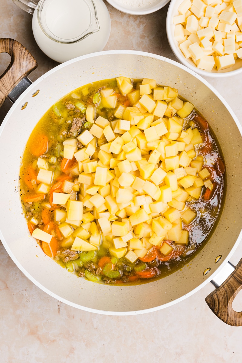 Potatoes being added to a large pot with veggies and broth.