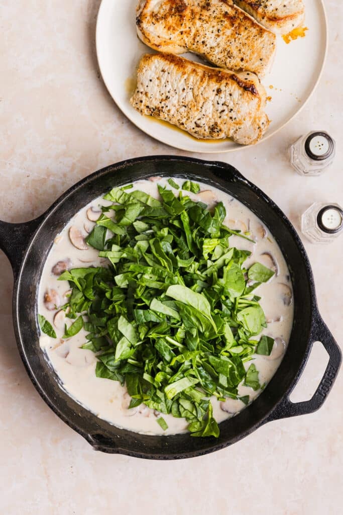 Spinach being added to a pan with a creamy sauce and mushrooms.