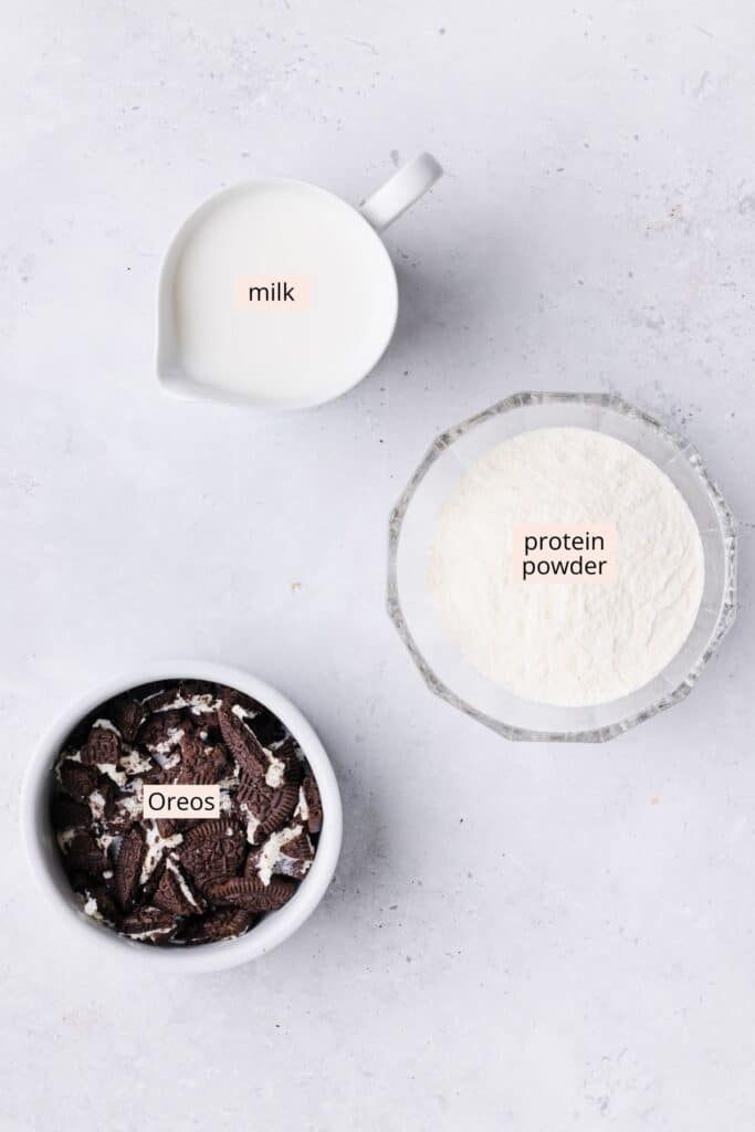 Protein powder, milk and oreos in bowls on a countertop with labels.