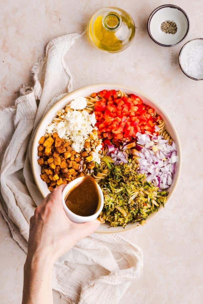 Balsamic dressing being poured onto pasta salad with whole wheat pasta, brussels sprouts, feta cheese, peppers, butternut squash and onions in a bowl.