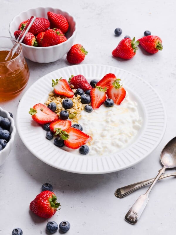 Cottage cheese, blueberries, strawberries and granola in a white bowl with a drizzle of honey.