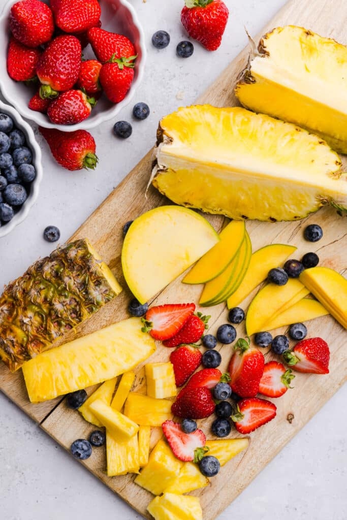 Pineapple, mango, strawberries and blueberries being sliced on a wooden cutting board for cottage cheese fruit bowls.