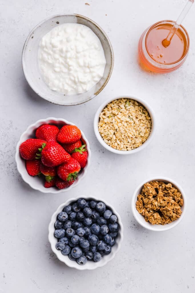 Cottage cheese, strawberries, blueberries, honey and granola in small bowls on a countertop.