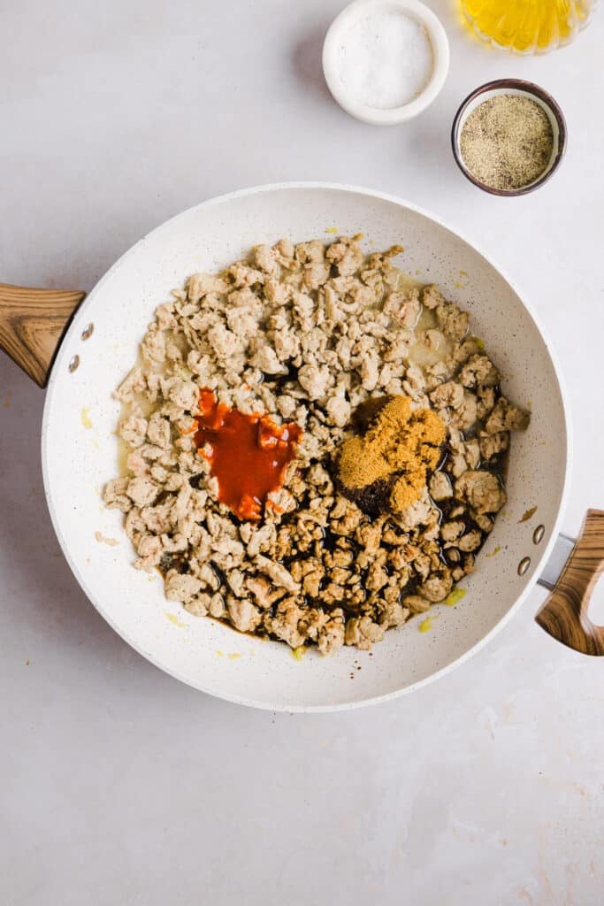 Spices and sriracha being added to ground chicken in a pan.