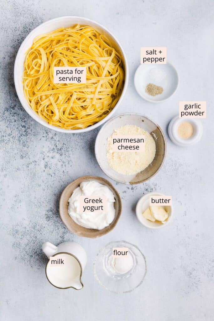 Ingredients for Greek yogurt alfredo sauce like Greek yogurt, parmesan cheese and butter on a countertop with labels.