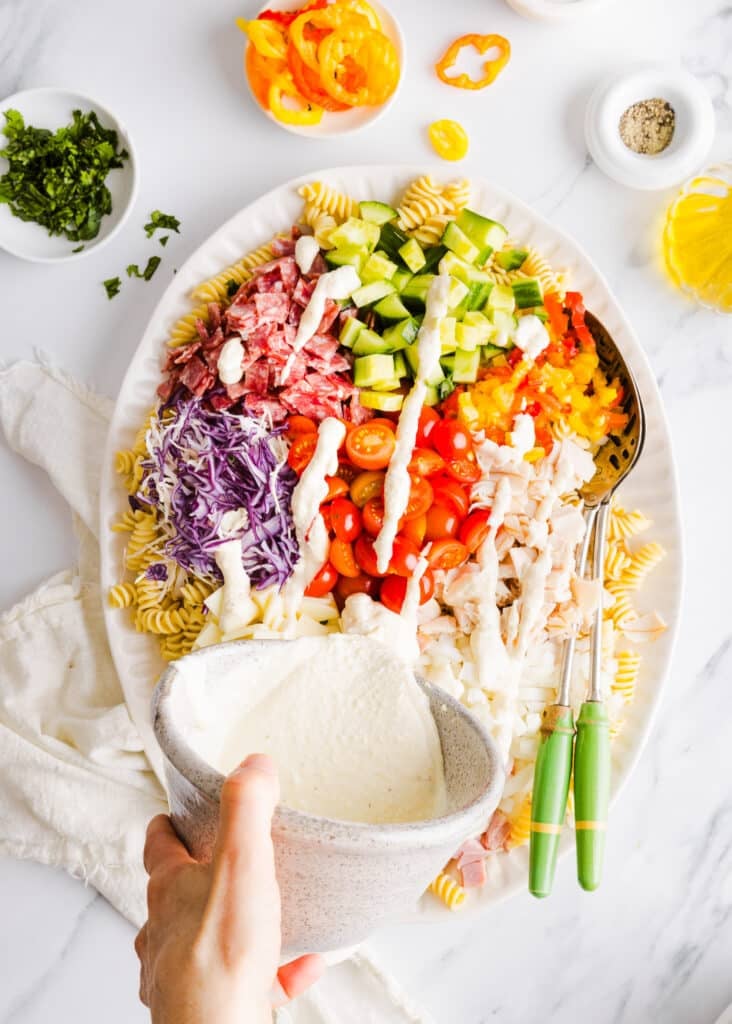 Grinder pasta salad dressing being poured onto a platter full of ingredients.