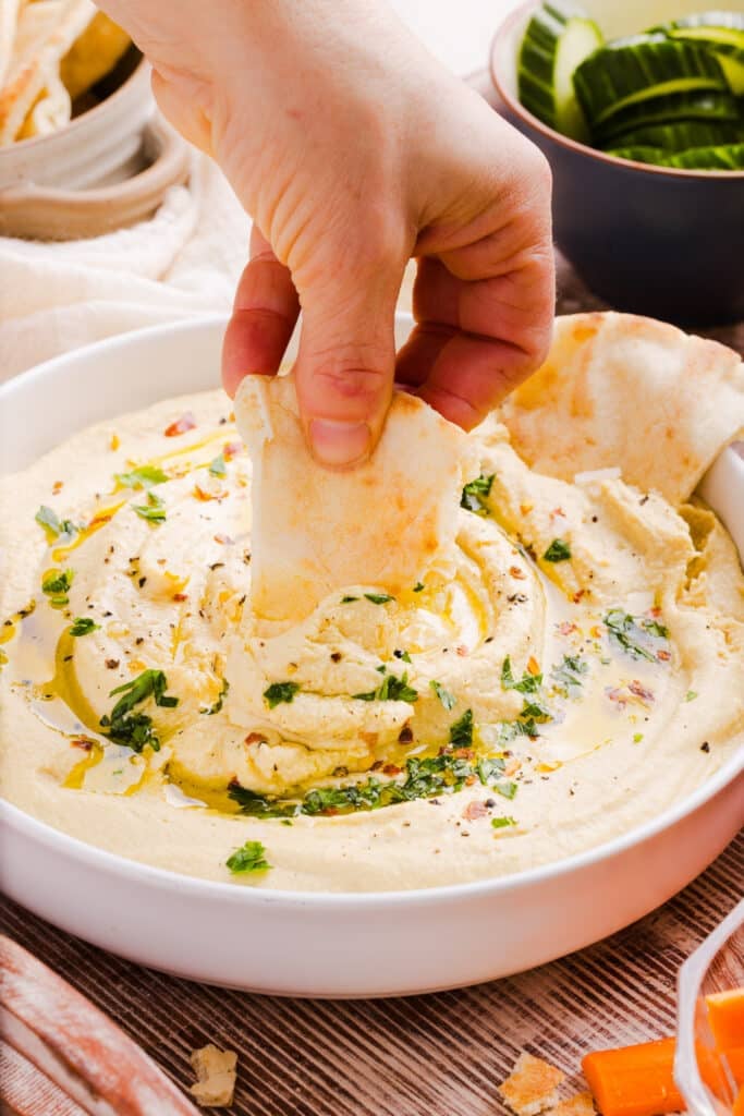A close shot of high protein hummus in a shallow bowl with olive oil, parsley and pita bread being dipped.