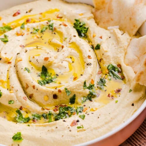 A close up shot of high protein hummus in a shallow bowl with olive oil, parsley and pita bread being dipped.