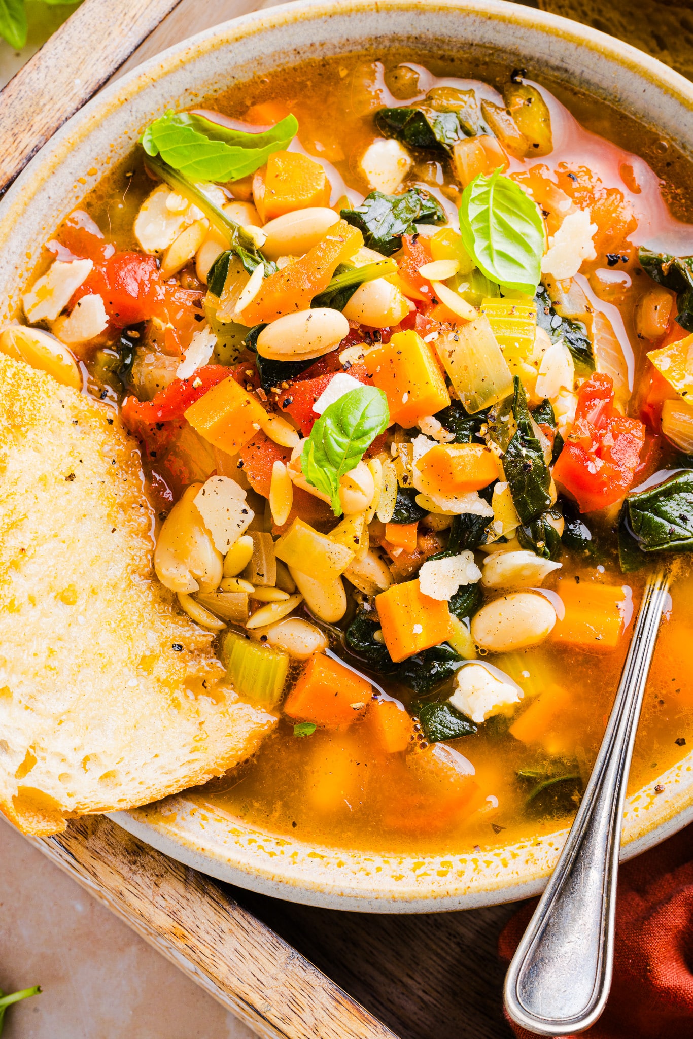 Vegetable orzo soup in a bowl with a piece of bread and a spoon.