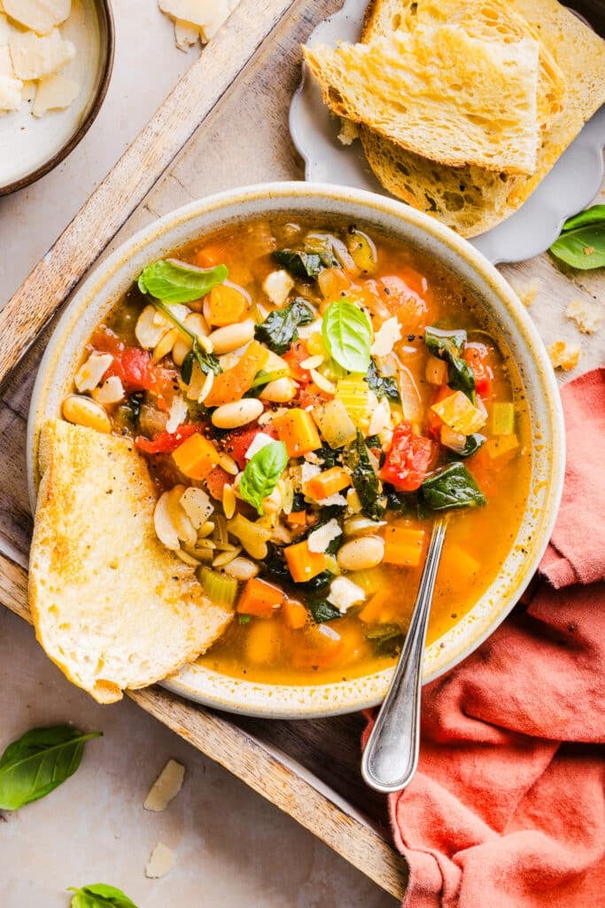 Vegetable orzo soup in a bowl with a piece of bread and a spoon.