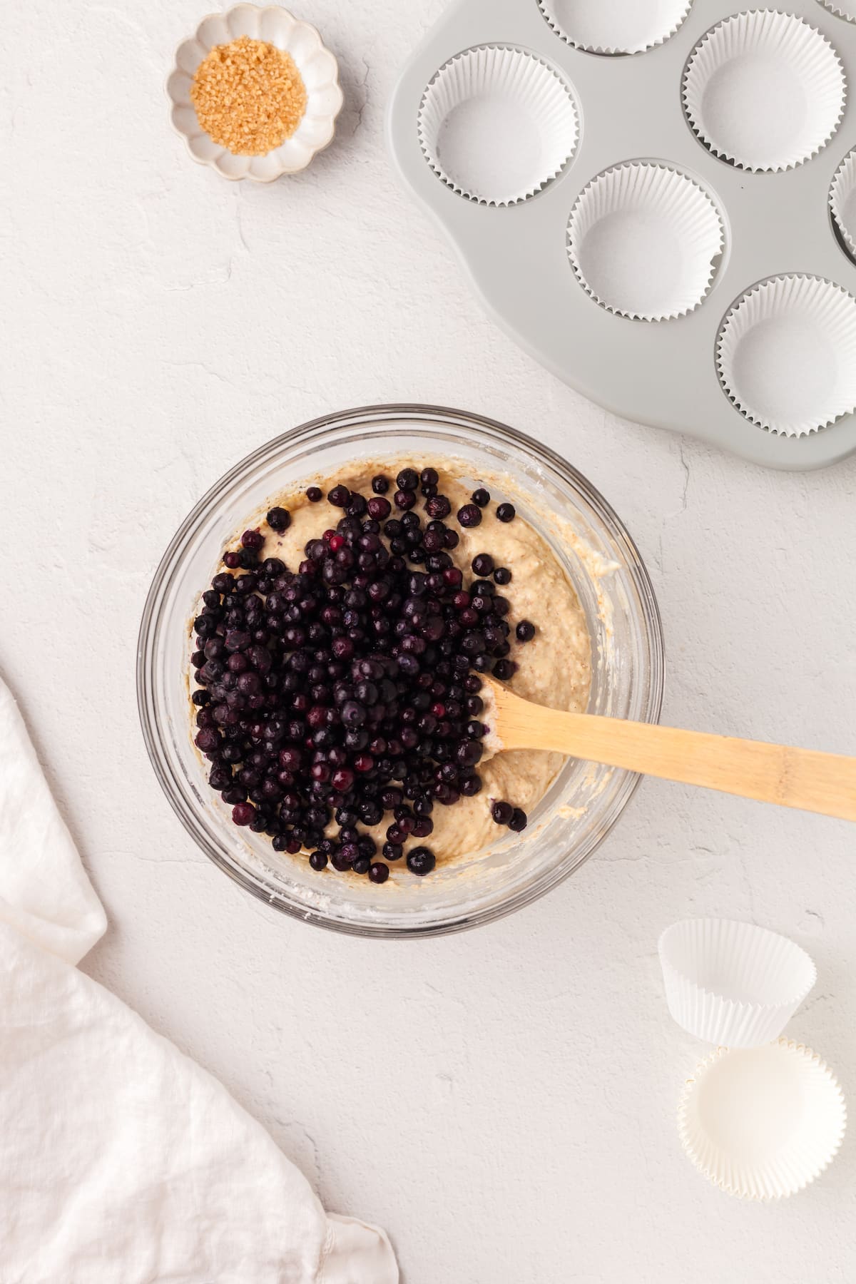 Frozen blueberries being added to muffin batter in a glass bowl.