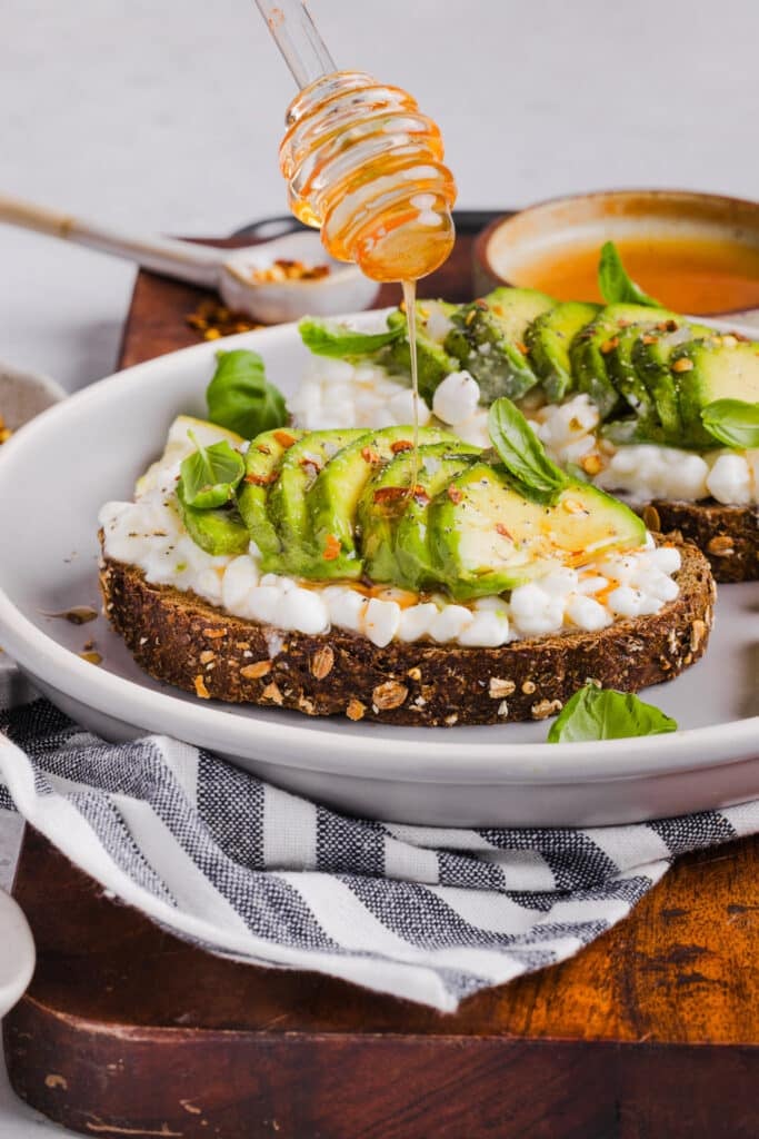 Hot honey being drizzled onto whole grain toast with cottage cheese.