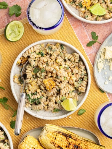 Elote pasta salad topped with cotija cheese and cilantro, on a tablecloth with margaritas and limes for serving.