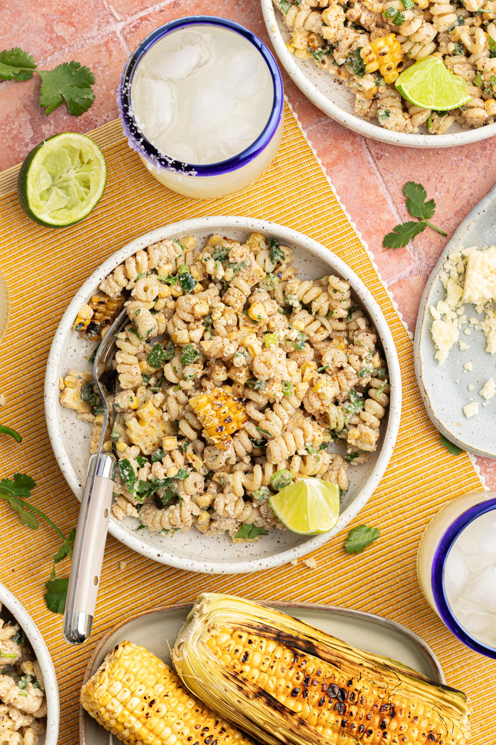 Elote pasta salad topped with cotija cheese and cilantro, on a tablecloth with margaritas and limes for serving.