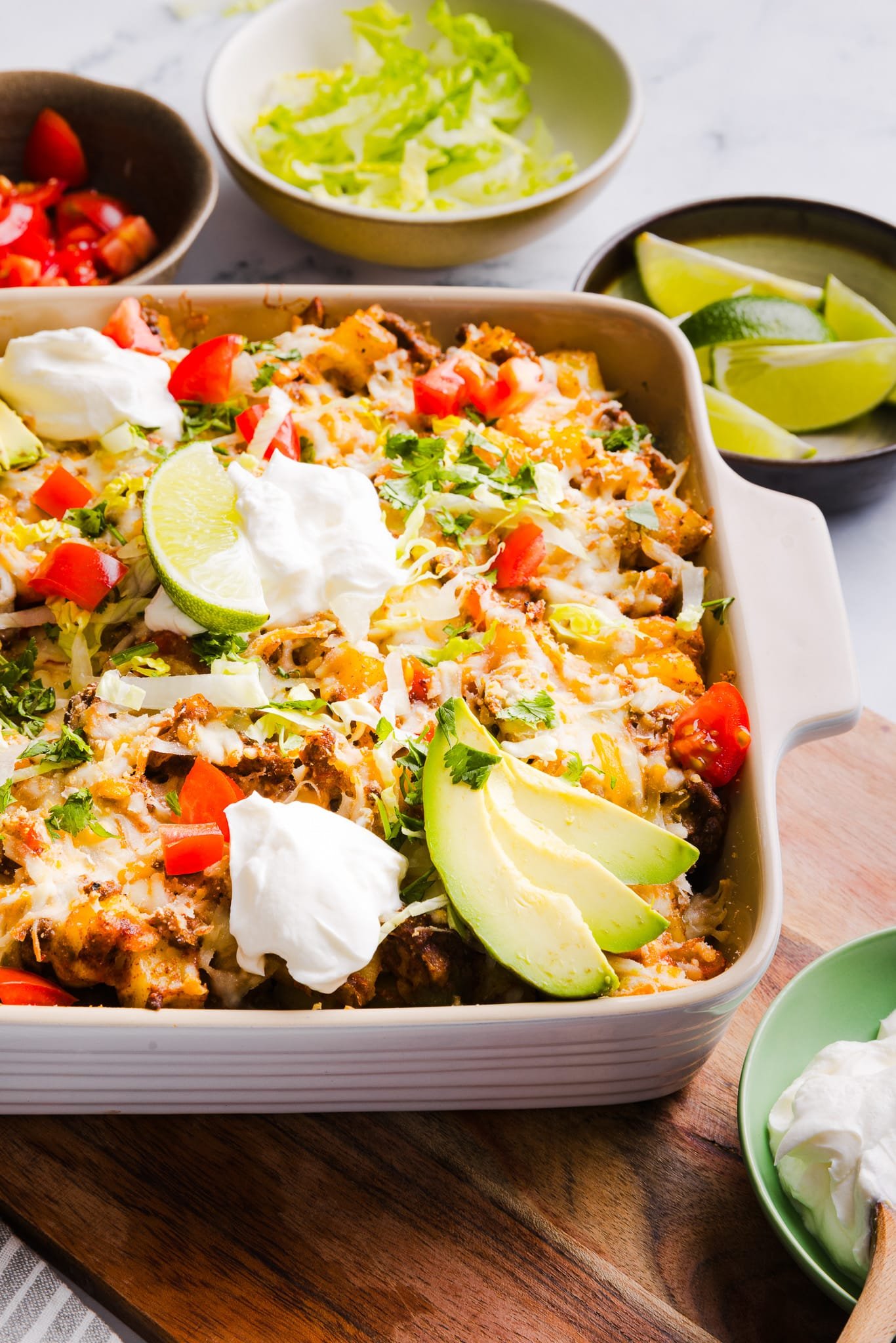 Taco potato casserole baked in a dish and topped with shredded lettuce, cheese, sour cream, avocado, lime and tomatoes.