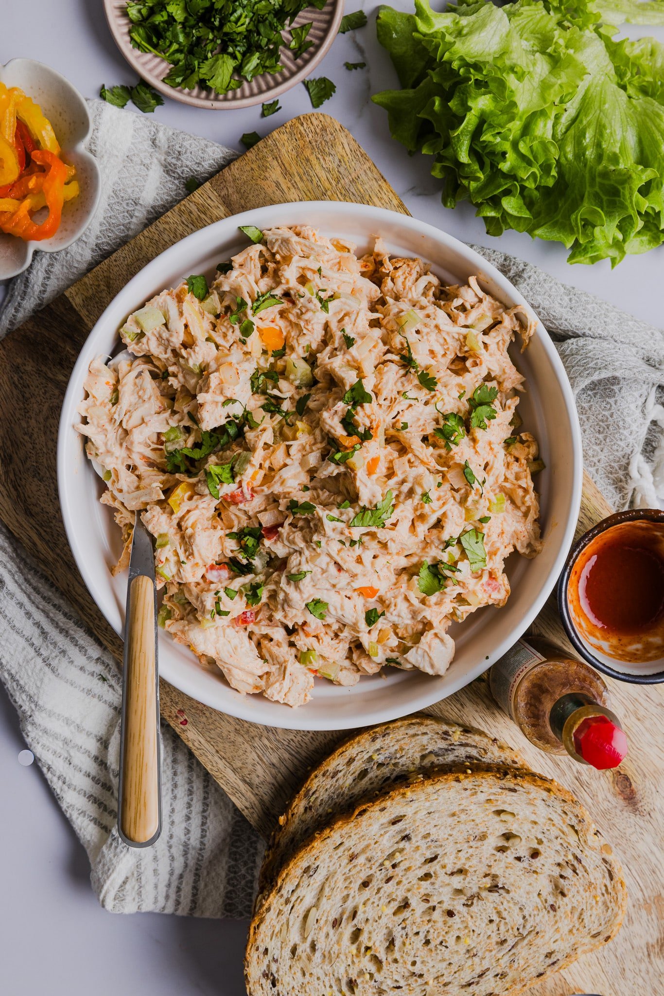 A bowl of chicken salad with lettuce, bread, and peppers next to it.