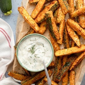 Air fryer zucchini fries on parchment paper with a bowl of Greek yogurt dill dip.