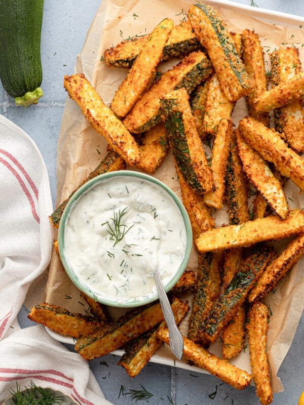Air fryer zucchini fries on parchment paper with a bowl of Greek yogurt dill dip.