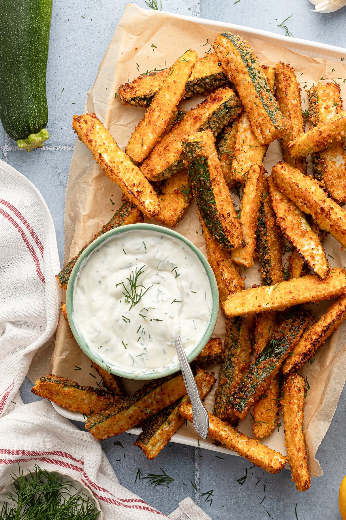 These crispy, air fryer zucchini fries with tangy Greek yogurt dill sauce are the perfect healthier alternative to traditional fries. Air fryer zucchini fries on parchment paper with a bowl of Greek yogurt dill dip.