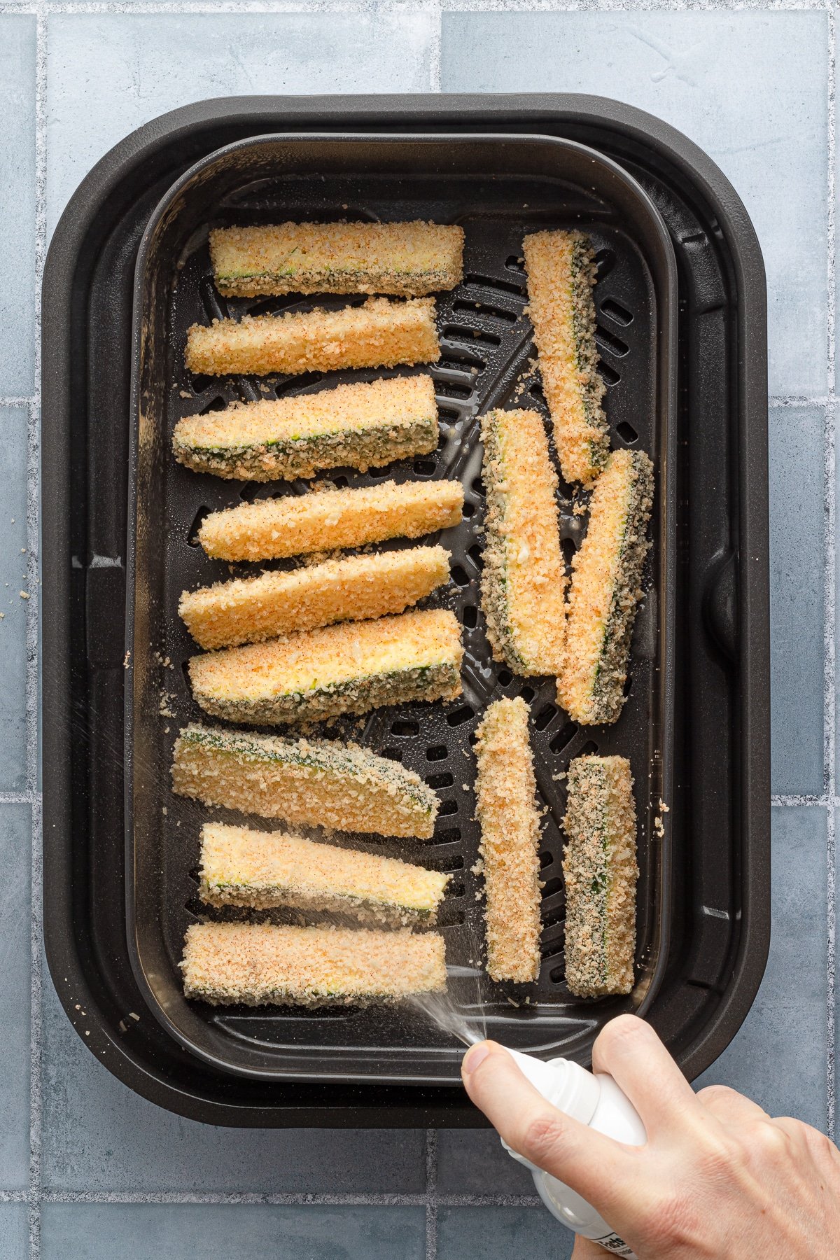 Spraying breaded zucchini spears with non-stick cooking spray in an air fryer.