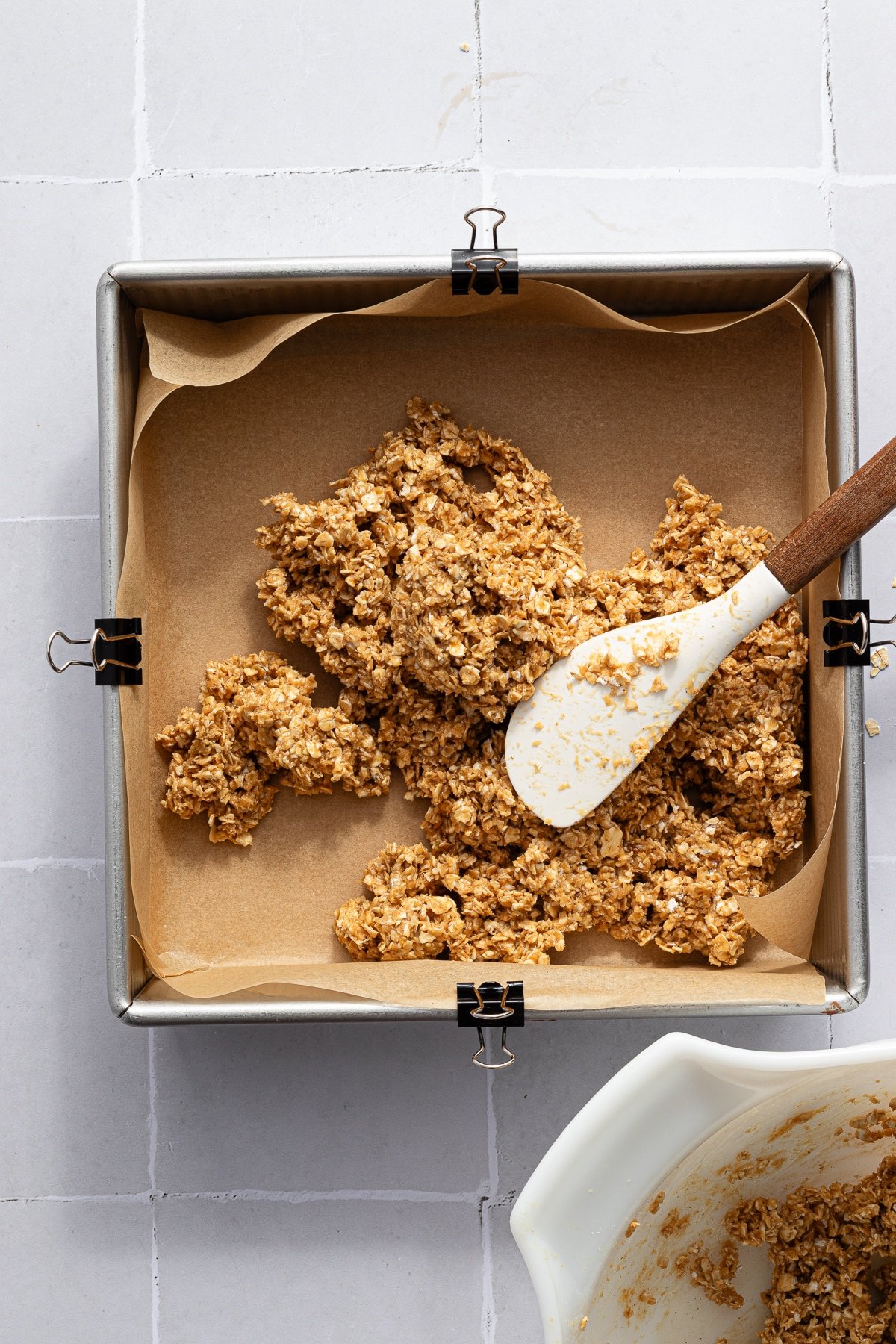 An oat mixture being added to a parchment paper lined square dish for chocolate oat bars.