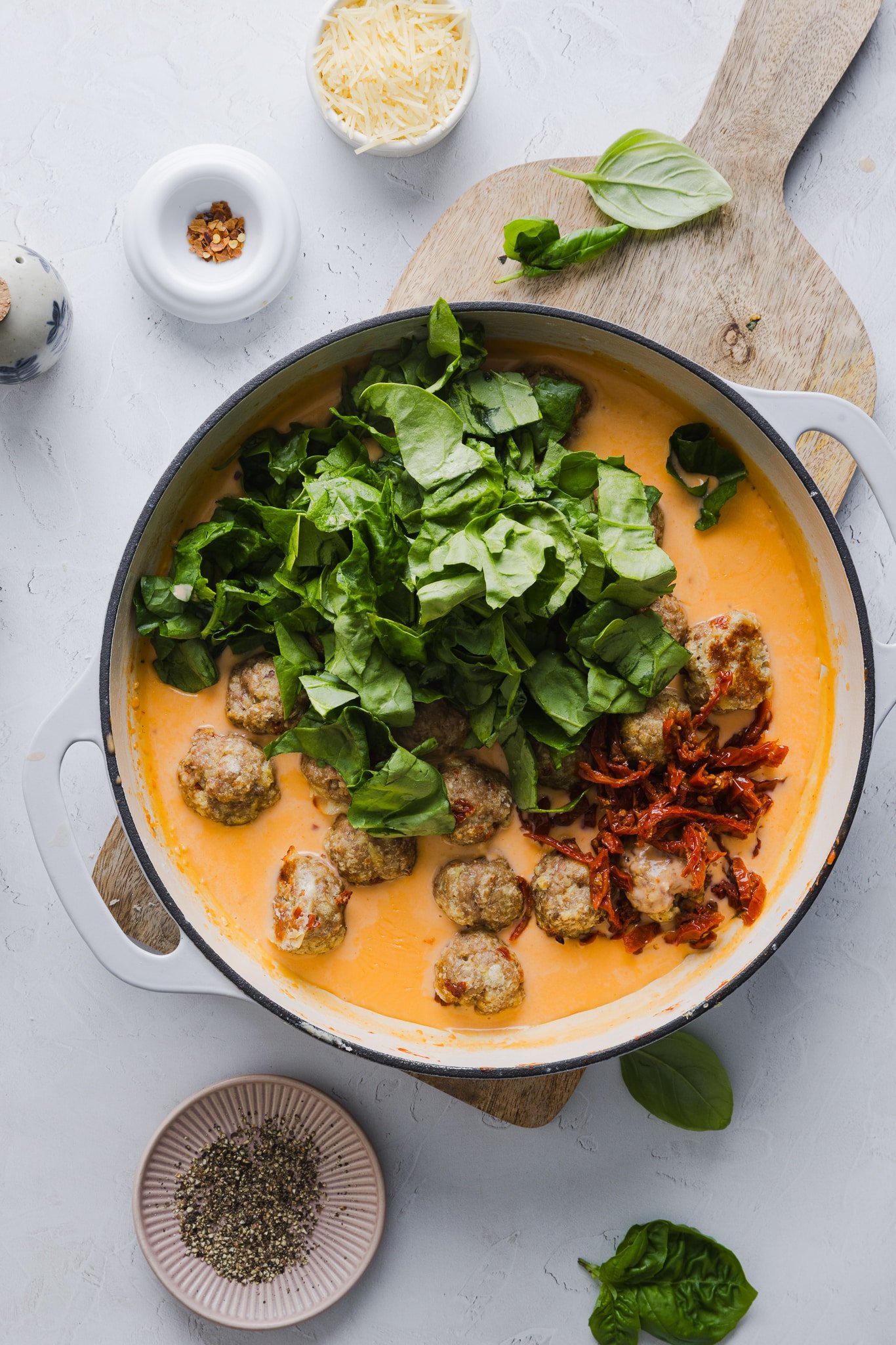 Chicken meatballs, sun-dried tomatoes and spinach being added to a pan with creamy sauce.