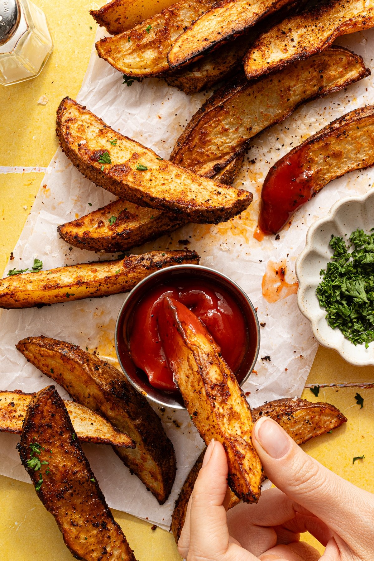Overhead image of a hand dipping a potato wedge in ketchup.