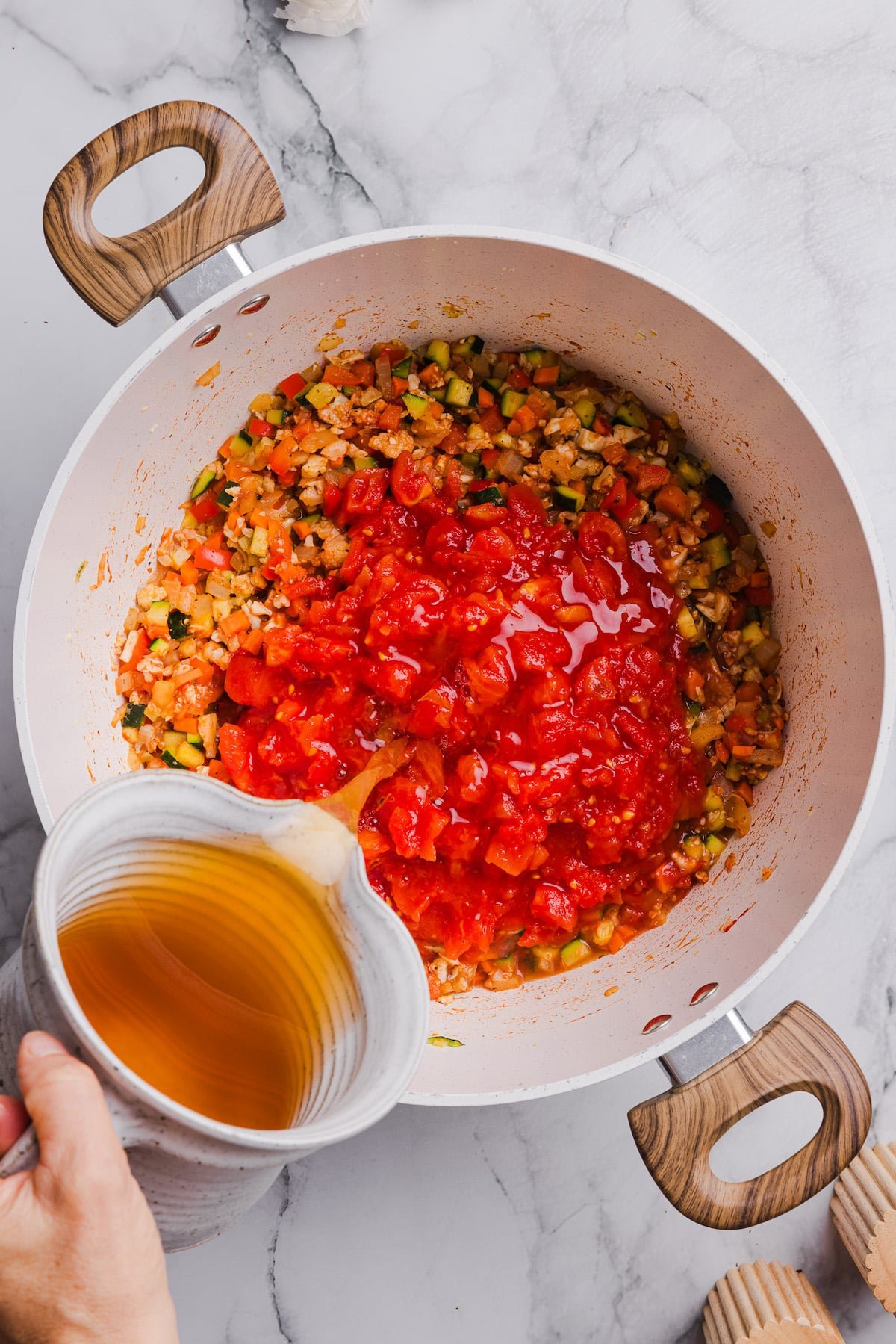 Crushed tomatoes and broth being added to sautéed veggies in a pot.