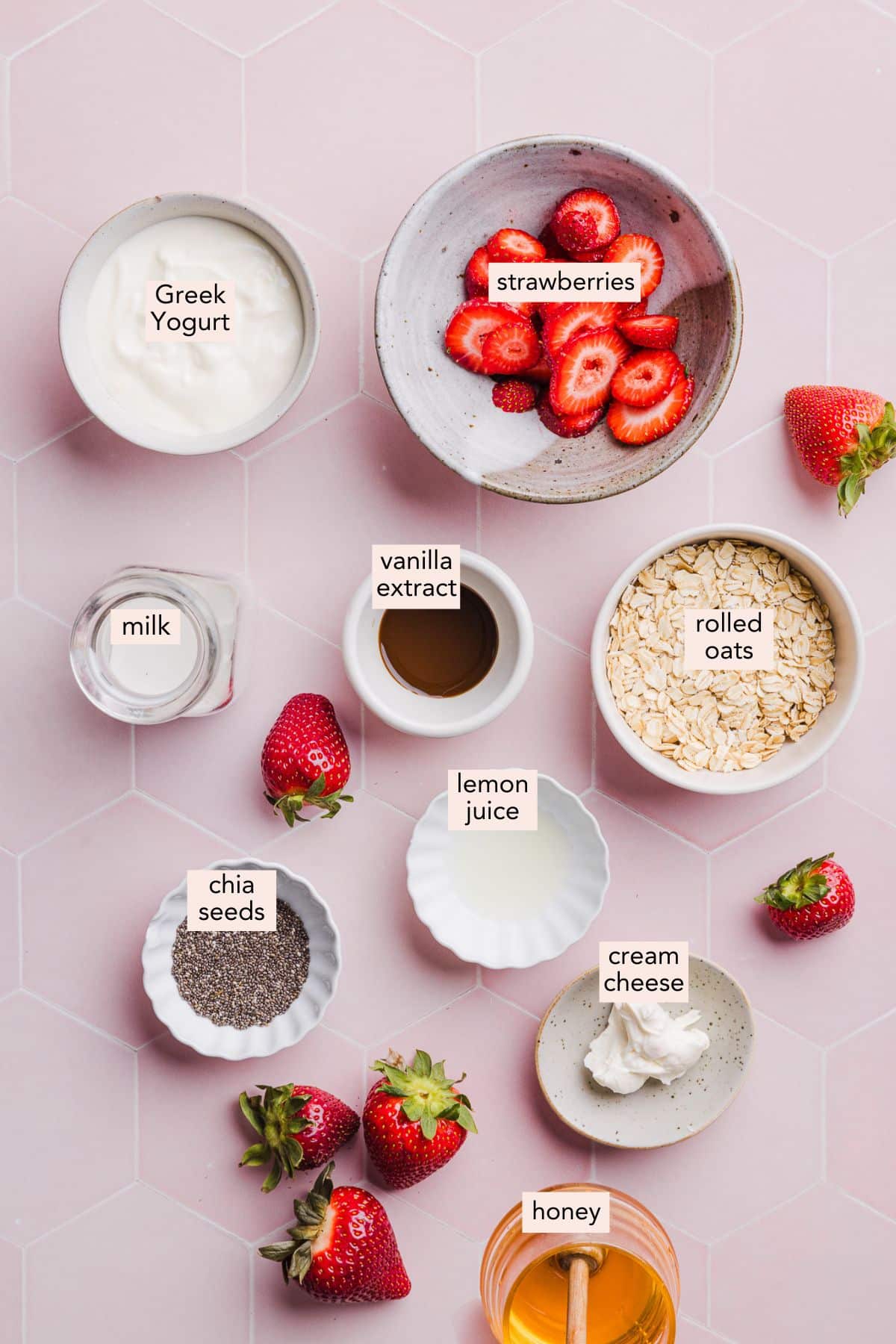 Ingredients for strawberry cheesecake overnight oats on a pink countertop with labels.