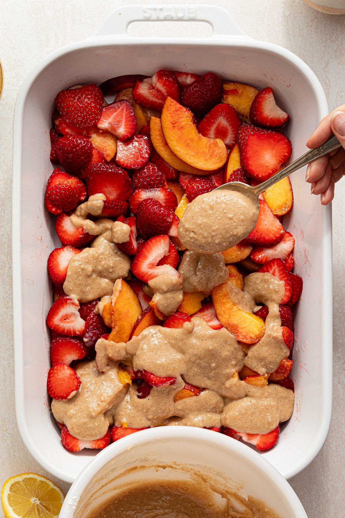Strawberry peach cobbler batter being spooned on strawberries and peaches in a white baking dish.