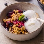 A serving of apple blueberry crumble in a bowl with two scoops of vanilla ice cream.