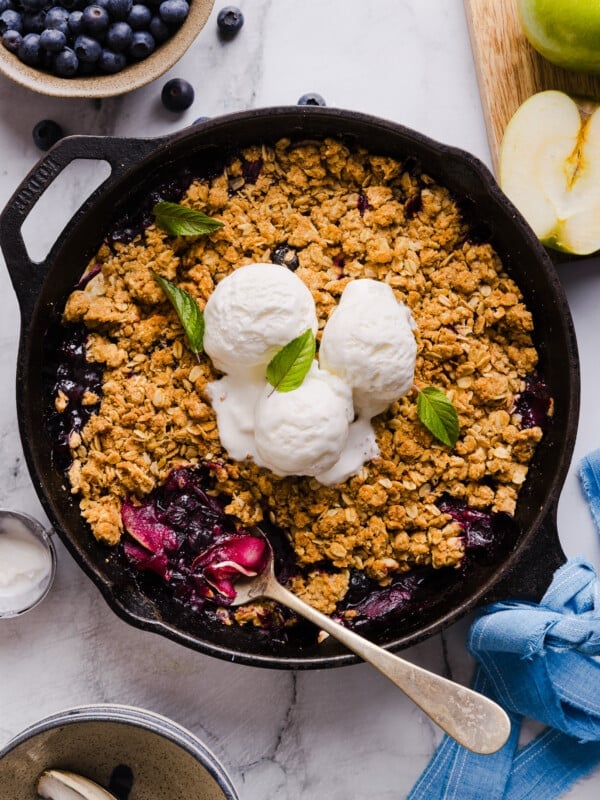 Apple blueberry crumble in a skillet, topped with three scoops of vanilla ice cream.
