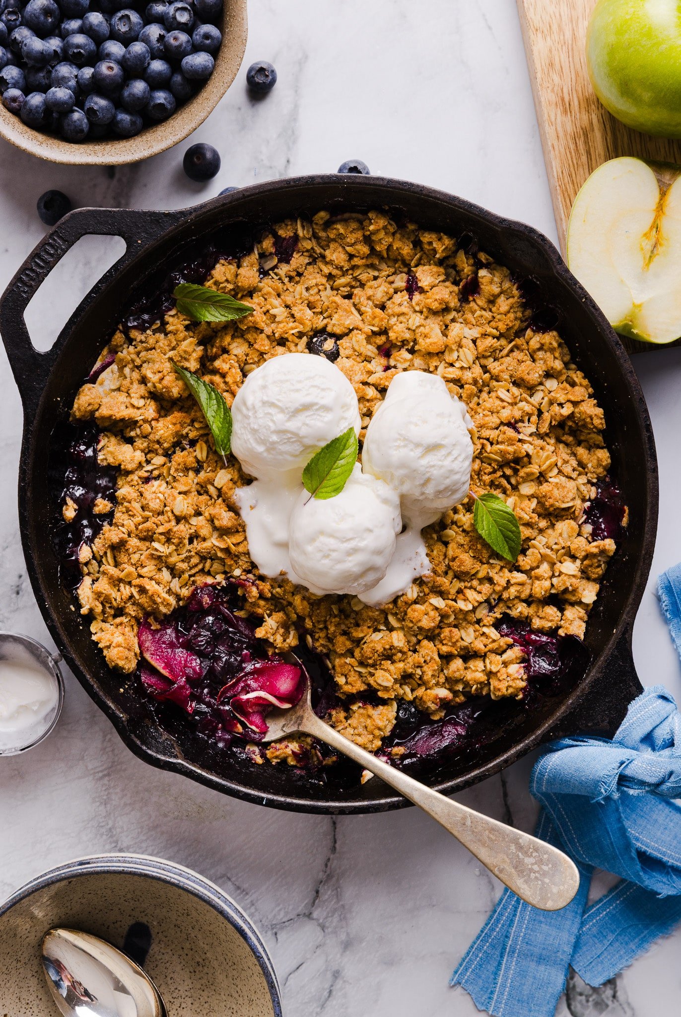 Apple blueberry crumble in a skillet, topped with three scoops of vanilla ice cream.